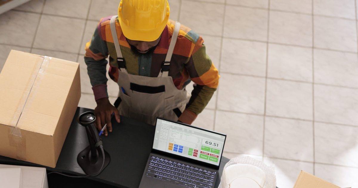 Top view of diverse team discussing customer order preparing packages for delivery in warehouse. African american employee wearing industrial overall and helmet working at goods quality control